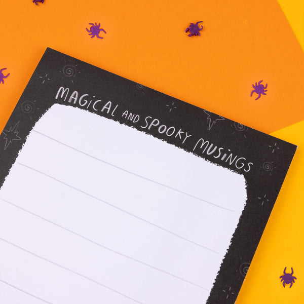 The 'Magical and Spooky Musings' notepad by Katie Abey features a black border with star and swirl accents, topped with hand-drawn text. Ideal for writing down your weird and wonderful thoughts, with illustrations of a pumpkin, mushroom, and bat along the bottom.