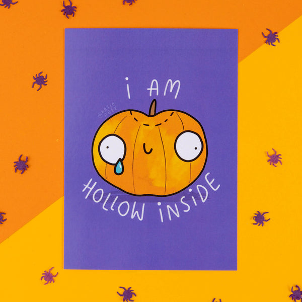 A whimsical Halloween-themed postcard by Katie Abey featuring a sad, cartoonish pumpkin with large, round eyes and a tear falling down its face. The pumpkin has the phrase "I am hollow inside" written around it in white text on a vibrant purple background. The card is placed on a bright orange surface decorated with small purple spider confetti, giving off playful, spooky vibes. Katie Abey is an illustrator and small business that encourages people to embrace their weirdness.