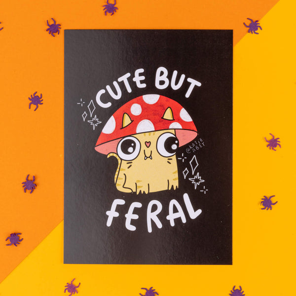 Katie Abey's 'Cute but Feral' postcard featuring an adorable cartoon cat wearing a red mushroom hat with white spots. The cat has big, round eyes and a quirky expression. The design encourages embracing weirdness with a playful and sassy tone. Displayed on an orange background scattered with small purple spiders, perfect for a spooky, fun vibe. Ideal for gifting or decorating. Created by Katie Abey, a small business illustrator inspiring you to be unapologetically yourself.