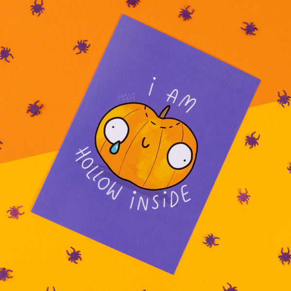 A whimsical Halloween-themed postcard by Katie Abey featuring a sad, cartoonish pumpkin with large, round eyes and a tear falling down its face. The pumpkin has the phrase "I am hollow inside" written around it in white text on a vibrant purple background. The card is placed on a bright orange surface decorated with small purple spider confetti, giving off playful, spooky vibes. Katie Abey is an illustrator and small business that encourages people to embrace their weirdness.