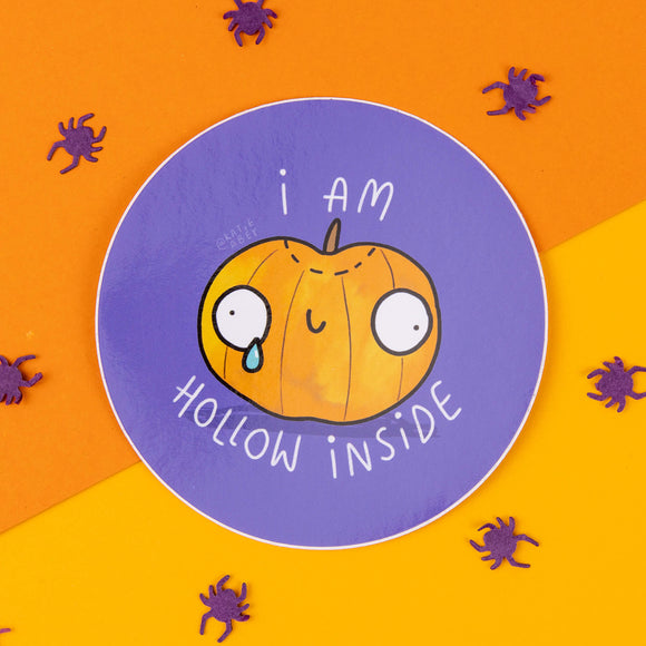 This circular sticker by Katie Abey features a cute cartoon pumpkin with a sad, teary expression and the words "I Am Hollow Inside" in playful white text. The sticker has a vibrant purple background, contrasting with the orange pumpkin. The brand's quirky and weirdly wonderful style shines through, making it a perfect addition to your Halloween-themed gifts. Purple spider confetti surrounds the sticker, adding to the spooky vibes. Katie Abey is a UK illustrator who celebrates embracing your weirdness.