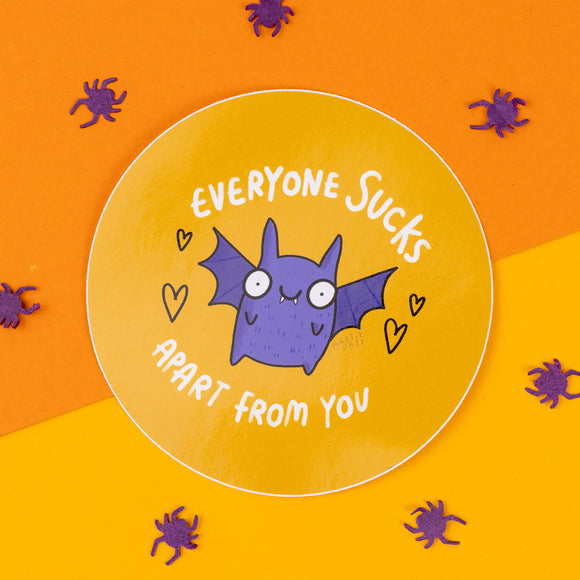 A round, bright orange and yellow sticker featuring a cute purple bat illustration by Katie Abey. The bat has wide, comical eyes and small wings. The text around the bat reads, "Everyone sucks apart from you," making this an amusing and sassy gift option. Small purple paper spiders are scattered around the sticker for a spooky, Halloween-themed vibe. Katie Abey, an illustrator and small business, encourages embracing your weirdness through fun, quirky designs like this one. Perfect for gifting!