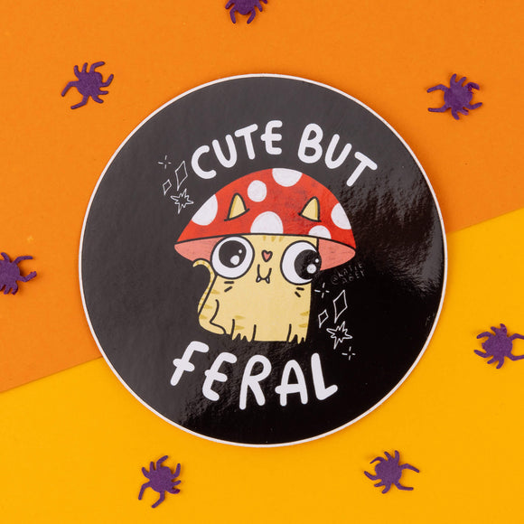 Round sticker from Katie Abey featuring an illustrated cartoon cat with wide eyes wearing a red and white spotted mushroom hat. The caption reads "Cute But Feral" in bold white letters. Surrounded by a bright orange and yellow background with scattered purple spider confetti. Katie Abey is an illustrator and small business encouraging you to embrace your weirdness with quirky, humorous designs. Ideal for adding a bit of fun and personality to your laptop, notebook, or gifts.