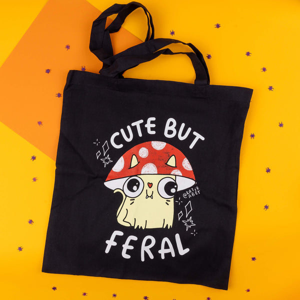 A black tote bag by Katie Abey, featuring an illustration of a cute, wide-eyed yellow cat with fangs under a red and white spotted mushroom. The text above reads "Cute But Feral" in bold white letters. The background is vibrant orange with small scattered purple bug-shaped confetti. Katie Abey is a UK-based illustrator encouraging embracing your weird side with quirky designs.