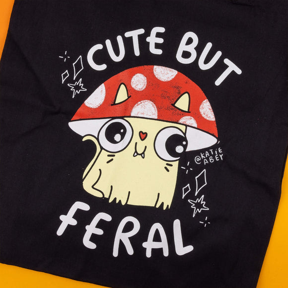 Close-up of a Katie Abey black tote bag, showcasing a yellow cat with fangs wearing a red and white mushroom hat. The words "Cute But Feral" appear in white, emphasising a playful, mischievous vibe. The bag is part of Katie Abey's collection, a UK-based illustrator promoting weirdness and fun through her quirky designs.