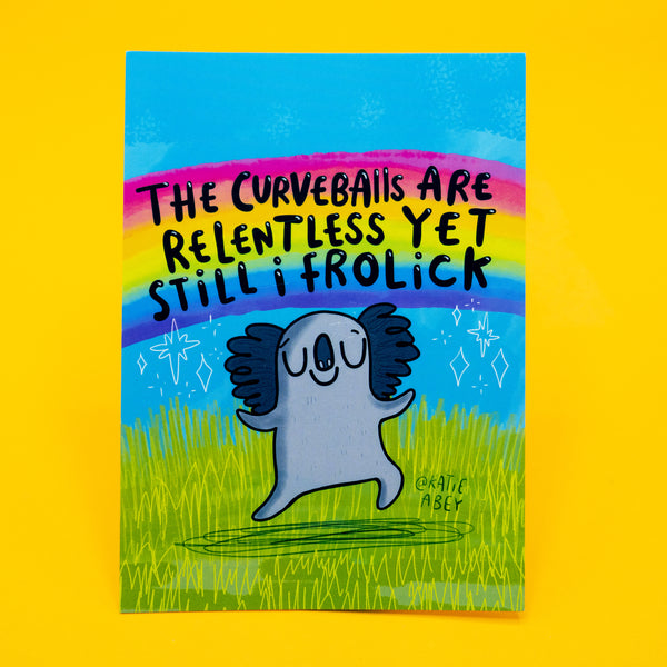 A6 postcard featuring a joyful cartoon koala leaping in a grassy field beneath the hand-lettered quote “The curveballs are relentless yet still I frolick.” A bold rainbow stretches across the blue sky, with sparkles and doodles adding to the whimsical feel. Printed on high-quality card, this uplifting design by Katie Abey encourages resilience and playful weirdness.