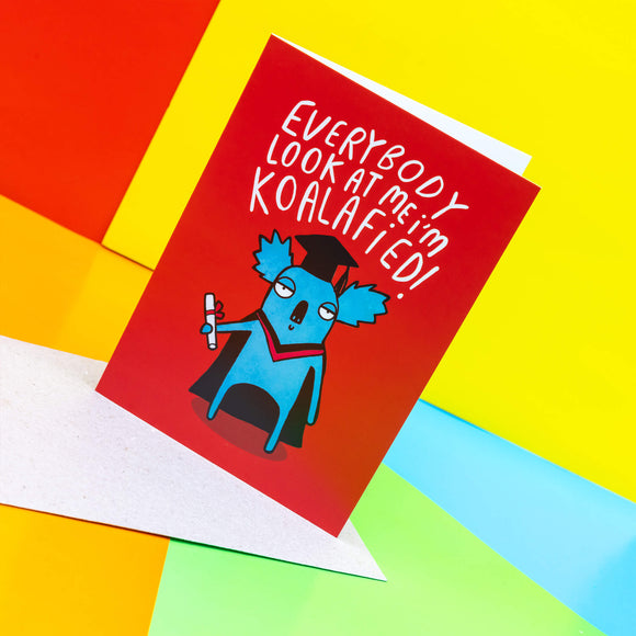 Everybody Look at me! I'm koalafied a6 greeting card, designed and printed by Katie Abey in the UK. The card cover is a red background with a koala wearing a graduation outfit and holding their certificate whilst looking smug. The text above the koala reads 'Everybody look at me I'm koalafied!' The card is stood up on the brown envelope it comes with on a multi coloured background.