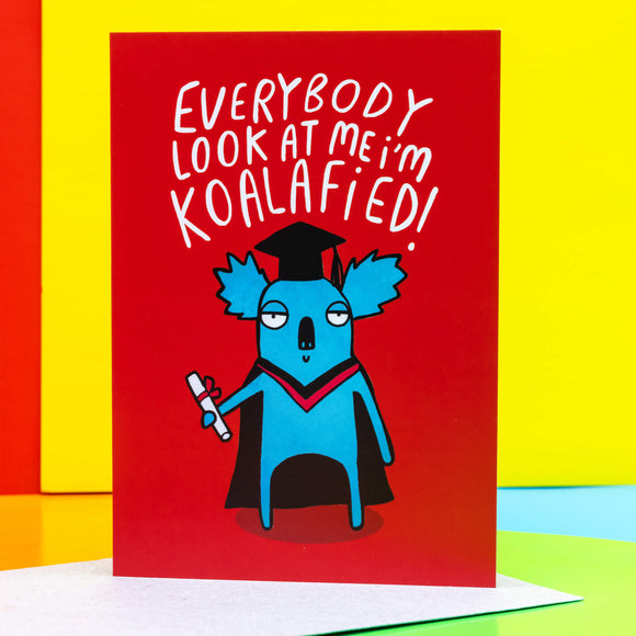 Everybody Look at me! I'm koalafied a6 greeting card, designed and printed by Katie Abey in the UK. The card cover is a red background with a koala wearing a graduation outfit and holding their certificate whilst looking smug. The text above the koala reads 'Everybody look at me I'm koalafied!' The card is stood up on the brown envelope it comes with on a multi coloured background.