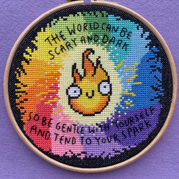 Close-up of the finished Tend To Your Spark cross stitch hoop by Katie Abey and Hoops & Crosses, featuring a cute flame surrounded by a rainbow burst and the words “The world can be scary and dark, so be gentle with yourself and tend to your spark.”