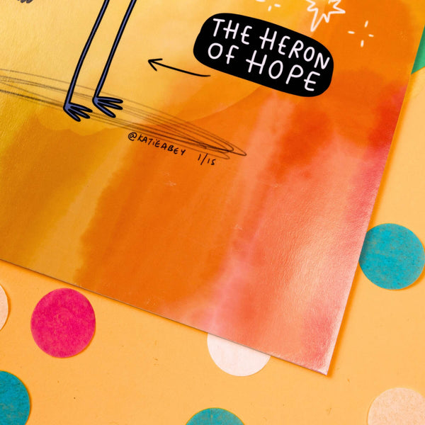 Heron Of Hope Art Print on a green, blue & yellow card with rainbow confetti. It is yellow, orange & red watercolour with a smiling heron wearing a rainbow sunflower hat with a speech bubble reading 'I just want you to know that you're on the right path and your existence makes a positive difference and so many beautiful moments lie ahead for you' with white sparkles and a black bubble reading 'the heron of hope'. Underneath is katie abey signature with 1/15.