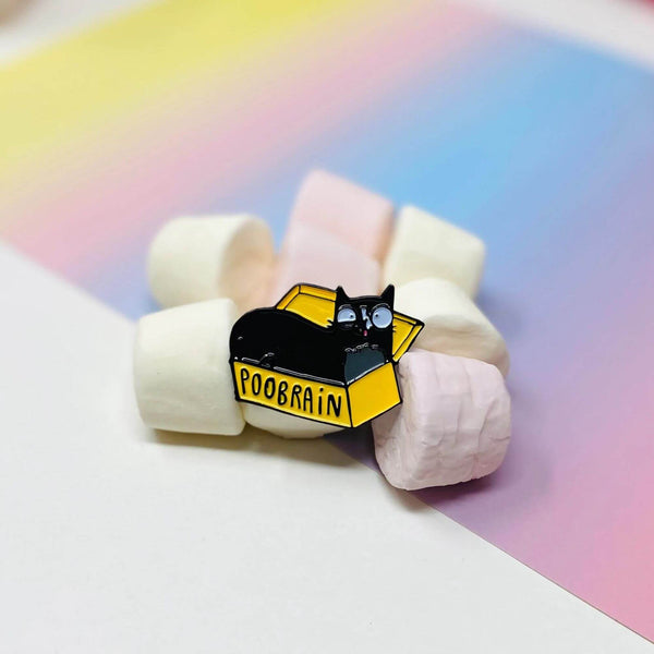 The Poobrain Cat Enamel Pin on some marshmallows. The enamel pin is of an open yellow box with a black cat inside with a silly expression. There is black text on the box that reads POOBRAIN. Designed by Katie Abey in the UK