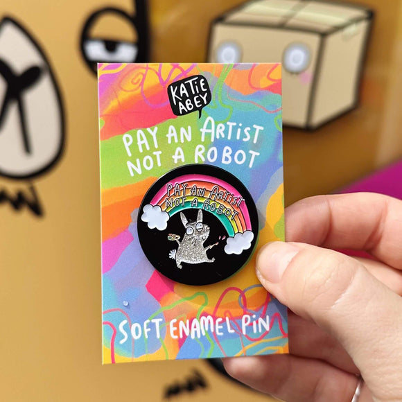 Pay an artist pin designed by Katie Abey in the UK. A black circle soft enamel pin with a sparkly grey rabbit wearing an artist hat and holding paint and brushes is smiling looking up at a rainbow reading 'pay an artist not a robot'. The pin is on a rainbow backing card with Katie Abey's logo at the top, then text that reads 'pay an artist not a robot' and bottom text that reads 'soft enamel pin'. The pin is being held up in front of the Katie Abey HQ.