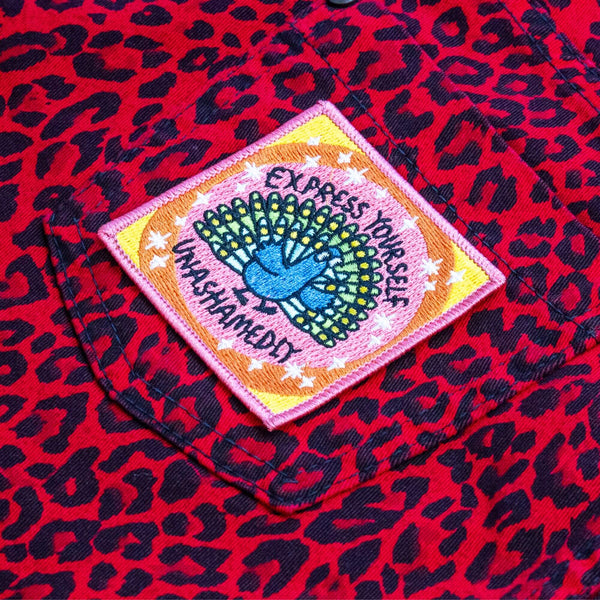 A patch with a peach background and a peacock illustration by Katie Abey in embroidery with Express yourself unashamedly written around it