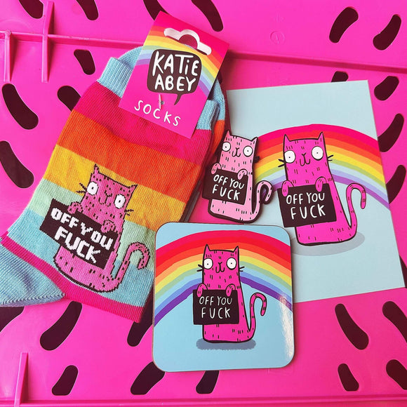 Contents of Off You F*ck Sweary Cat Bundle on pink background. Contents includes socks, a coaster, postcard and magnet. All products are light blue and rainbow coloured with a pink cat illustration holding a black sign sign with 'off you f*ck' written in white letters.