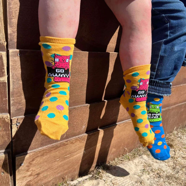  Yellow socks with blue and purple polka dots and pink cat illustration holding a black sign that reads 'go away'. The socks are being worn by child's feet.