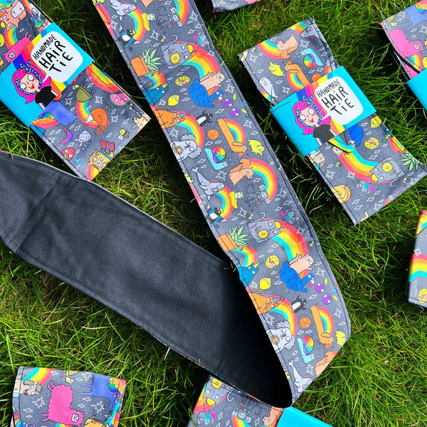 Multiple Positivity Print Hair Ties in its sky blue cardboard packaging laid on the grass. The light grey hair tie has illustrations of Katie Abey characters all over.