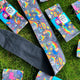 Multiple Positivity Print Hair Ties in its sky blue cardboard packaging laid on the grass. The light grey hair tie has illustrations of Katie Abey characters all over.