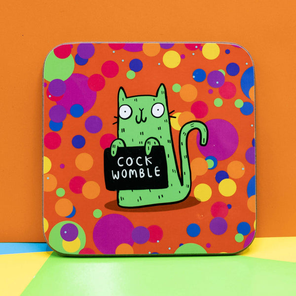 Sweary Womble Green Cat holding a sign saying, 'cock womble' on an orange background with multi coloured circles on a Coaster illustrated by Katie Abey