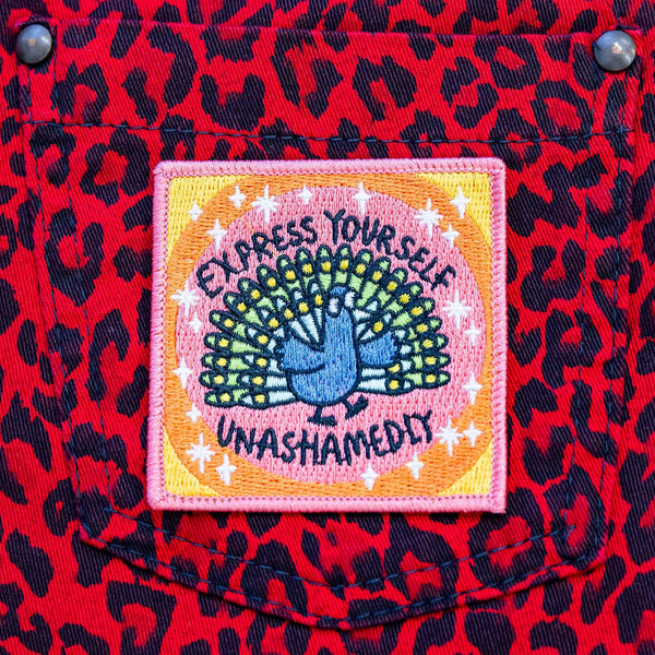 A patch with a peach background and a peacock illustration by Katie Abey in embroidery with Express yourself unashamedly written around it