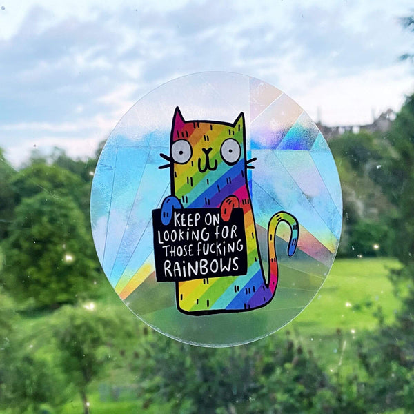 A see through rainbow making sun catching sticker that has a rainbow cat holding a sign saying keep on looking for those fucking rainbows