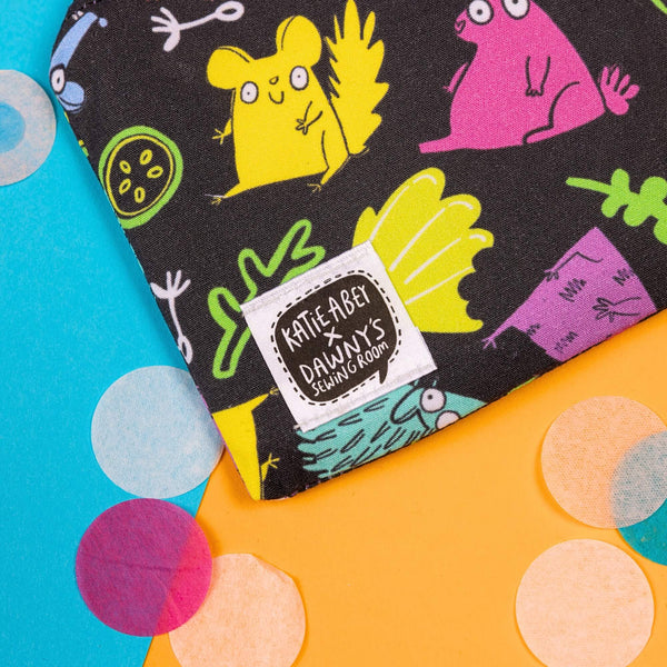 Close up of the Small Pets Coin Purse on yellow, blue and green card with rainbow circle confetti. The small black base coin purse has various colourful small pets such as chinchillas and rabbits with leaves and fruit all over, it has a black zip and a katie abey x dawny's sewing room tag in the bottom left corner.