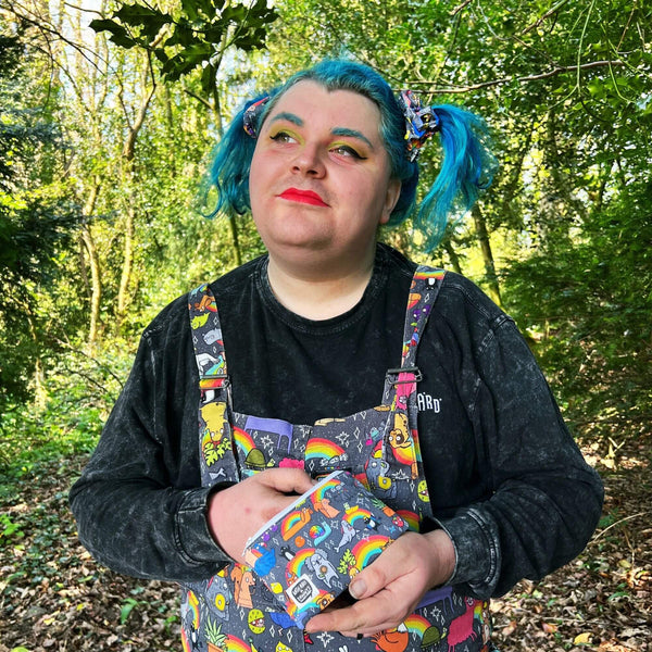 Jeff with blue hair and bright makeup is holding the Positivity Print Coin Purse in a woodland and is also wearing the matching dungarees