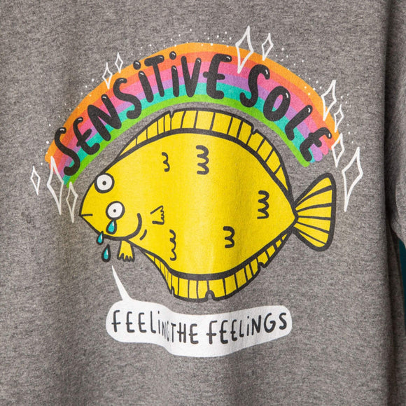 The grey t shirt has an illustration of a yellow sole with tears from it's eyes with a rainbow above it with the text sensitive sole written in front. There is a speech bubble from the sole's mouth that reads feeling the feelings
