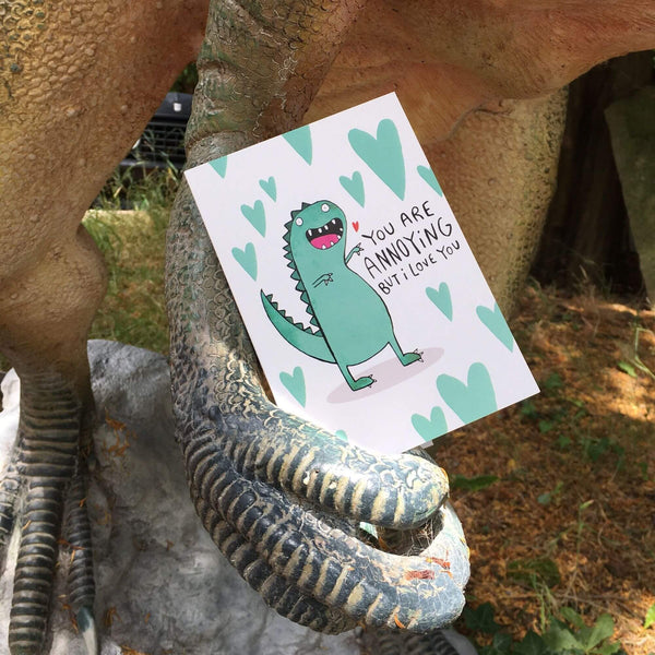 You're annoying but I love you a6 greeting card designed and printed by Katie Abey in the UK. The card is being held upright inside a dinosaur statue claw. The card front cover is a white background with a green t-rex style dinosaur smiling with multiple green hearts surrounding it and text reading 'You are annoying but I love you'.