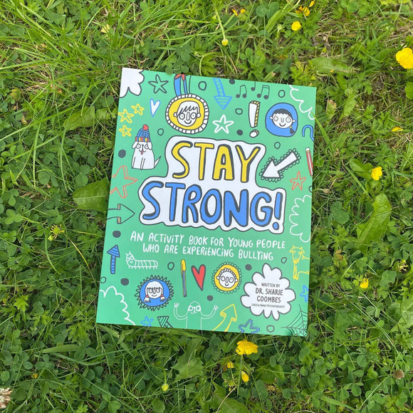 Stay Strong Book a collaboration with illustrations by Katie Abey and written by Dr. Sharie Coombes, Child & Family Psychotherapist with an introduction and notes for grown-ups. The book cover is a teal colour with various faces, symbols, animals, stars and stationary doodles bordering centre text that reads 'Stay Strong! An activity book for young people who are experiencing bullying'. The book is laying flat on a green grassy area.