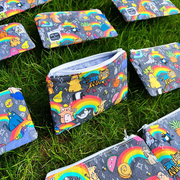 Multiple Positivity Print Coin Purses laid on grass. The light grey purse has Katie Abey character illustrations all over