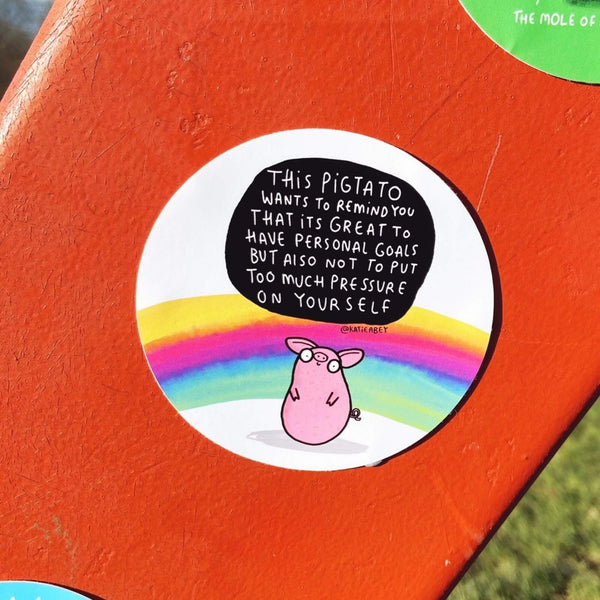 Circular Pigtato vinyl sticker for car windows, laptop, notebooks. Features a cute smiley pink pig illustration, on a white background with a rainbow, black speech bubble reads 'This pigtato wants to remind you that its great to have personal goals but also not to put too much pressure on yourself'