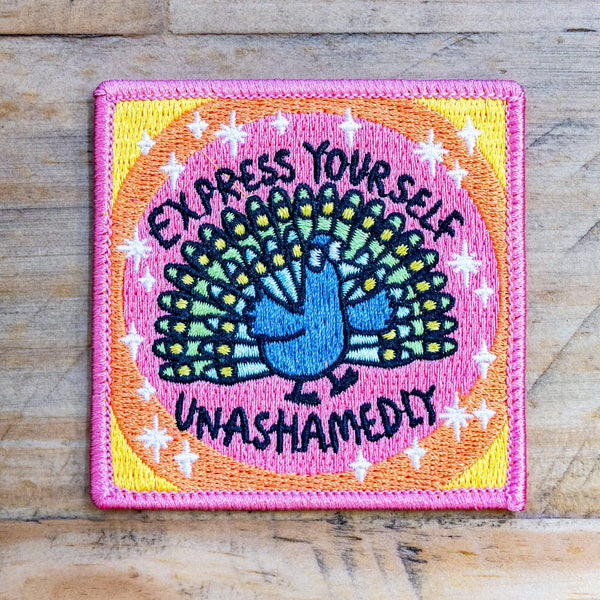 A patch with a peach background and a peacock illustration by Katie Abey in embroidery with Express yourself unashamedly written around it