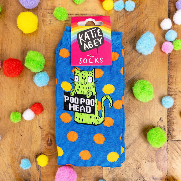 Blue socks on wooden background with fluffy rainbow balls sprinkled around. The blue socks have orange and yellow polka dots. Socks have lime greet cat illustration holding a black sign with 'poo poo head' written on in white letters.