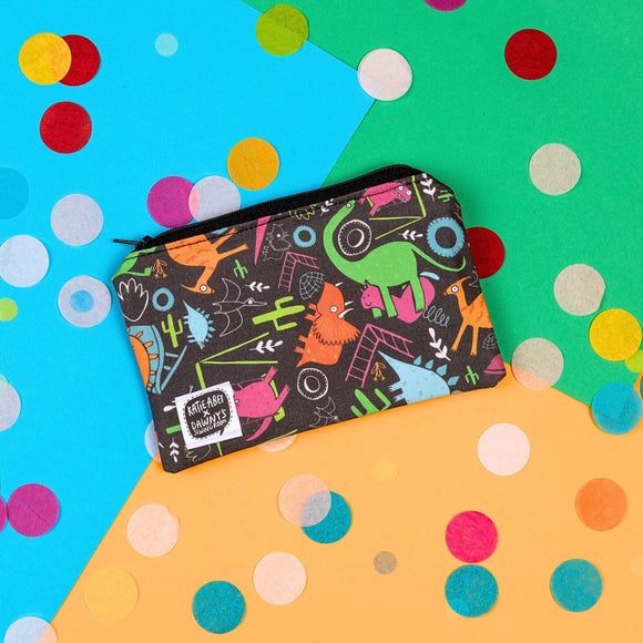 The Dinosaur Park Coin Purse on yellow, blue and green card with rainbow circle confetti. The black base coin purse has various colourful dinosaurs on park rides with traditional outdoor park climbing frames and toys, it has a black zip and a katie abey x dawny's sewing room tag in the bottom left corner.