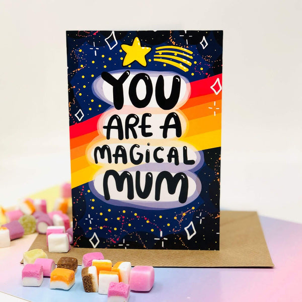 You are a magical mum a6 greeting card stood up on the brown envelope it comes with, pastel rainbow paper, dolly mixture sweets surrounding it on a white background. The front cover is a dark blue to black galaxy background with various red, white, yellow stars and sparkles, a red, orange, yellow gradient stripes going across the centre. The centre text reads 'You are a magical mum' with a yellow shooting star above it. Designed by Katie Abey in the UK.