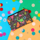 The Dinosaur Park Coin Purse on yellow, blue and green card with rainbow circle confetti. The black base coin purse has various colourful dinosaurs on park rides with traditional outdoor park climbing frames and toys and it has a black zip opening.