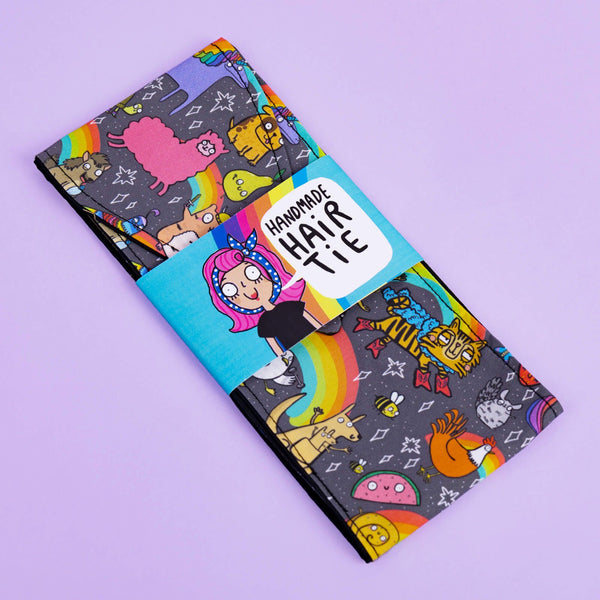 The Positivity Print Hair Tie in its sky blue cardboard packaging in front of a lilac backdrop. The light grey hair tie has illustrations of Katie Abey characters all over.