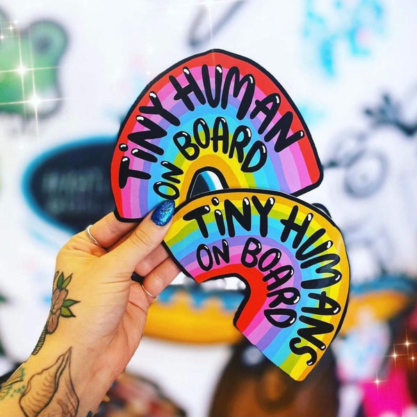 Tattooed hand with blue glittery nails holding two rainbow shaped vinyl stickers with black writing on, one reads 'tiny human on board' and the other 'tiny humans on board'. They are being held in front of a window with the sun shining in. Designed by Katie Abey in the UK.