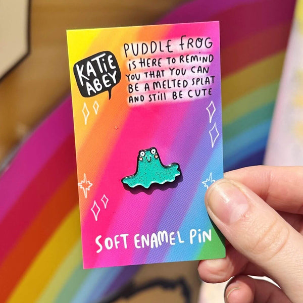 Puddle Frog Pin. A sparkly green frog shaped soft enamel pin with round white eyes, pink cheeks and cute smile. The pin is attached to rainbow coloured card with Katie Abey logo and writing that reads 'puddle frog is here to remind you that you can be a melted splat and still be cute'.