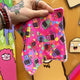Shot of reusable large makeup wipe, the wipe is a hot pink colour with various rainbow spots and cats holding black signs with rude words on.