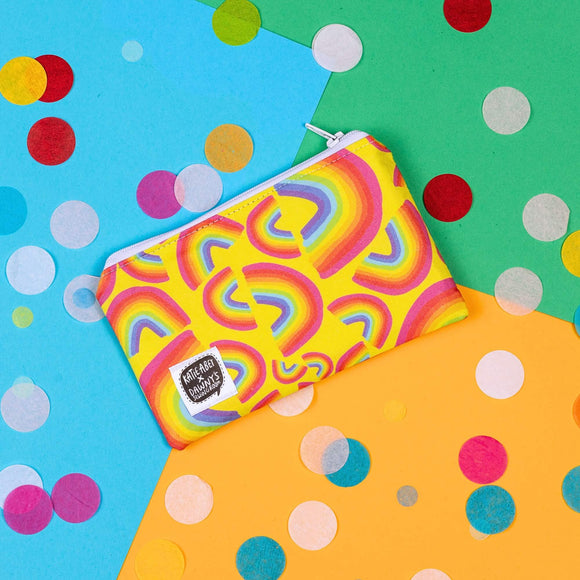 The Yellow Rainbow Coin Purse on yellow, blue and green card with rainbow circle confetti. The yellow base coin purse has rainbow arches in various sizes all over, it has a white zip and a katie abey x dawny's sewing room tag in the bottom left corner.