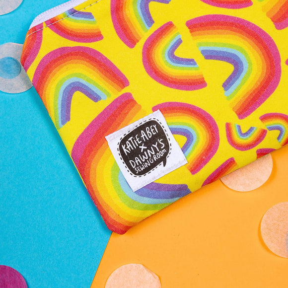 Close up of the Yellow Rainbow Coin Purse on yellow, blue and green card with rainbow circle confetti. The yellow base coin purse has rainbow arches in various sizes all over, it has a white zip and a katie abey x dawny's sewing room tag in the bottom left corner.