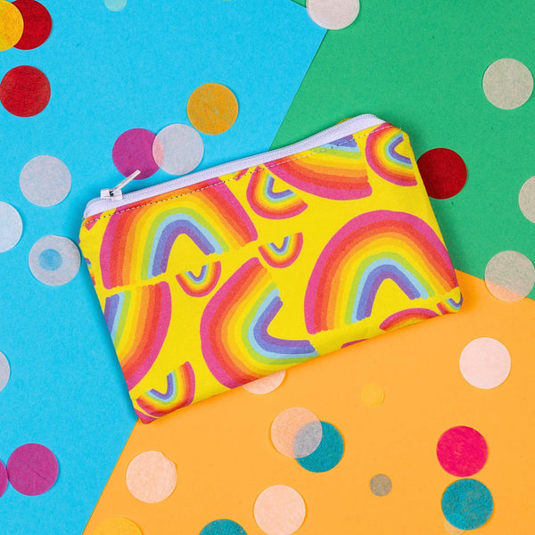 The Yellow Rainbow Coin Purse on yellow, blue and green card with rainbow circle confetti. The yellow base coin purse has rainbow arches in various sizes all over and it has a white zip.