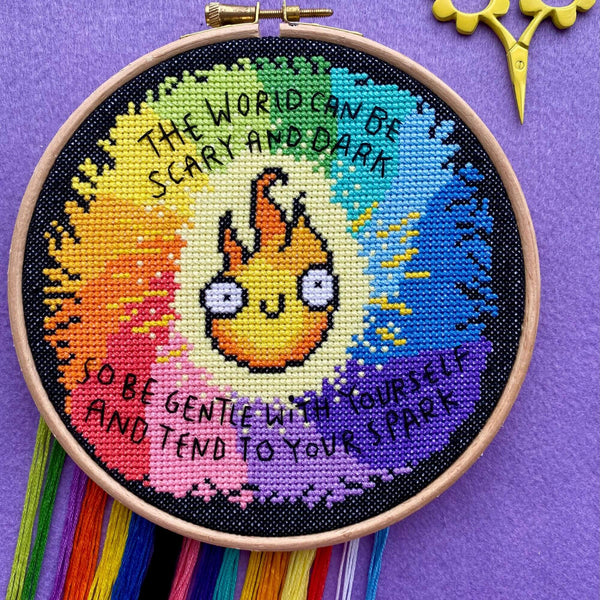  Finished rainbow cross stitch hoop with a flame character in the centre, photographed with yellow scissors and rainbow threads hanging from the bottom. Text reads “The world can be scary and dark so be gentle with yourself and tend to your spark.”
