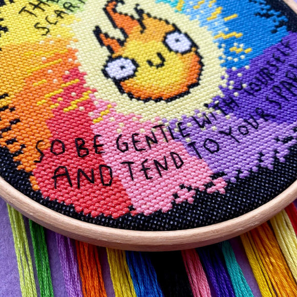 Close-up of the rainbow cross stitch with focus on the pink and red section and stitched words: “So be gentle with yourself and tend to your spark.” Threads dangle below.