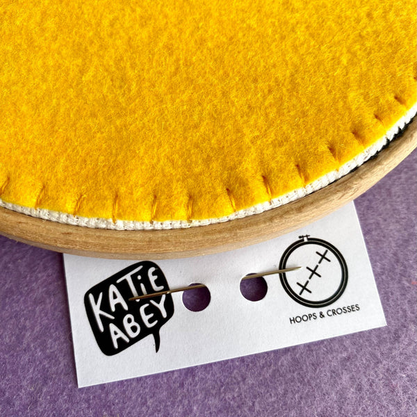 Back of the finished embroidery hoop showing yellow felt backing with Katie Abey and Hoops & Crosses branding card underneath.