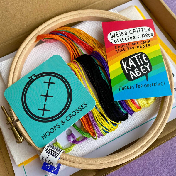 Unboxed cross stitch kit contents including embroidery hoop, DMC threads in rainbow colours, Aida fabric, and a colourful Katie Abey collector card. Hoops & Crosses branding visible on packaging.