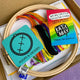 Unboxed cross stitch kit contents including embroidery hoop, DMC threads in rainbow colours, Aida fabric, and a colourful Katie Abey collector card. Hoops & Crosses branding visible on packaging.
