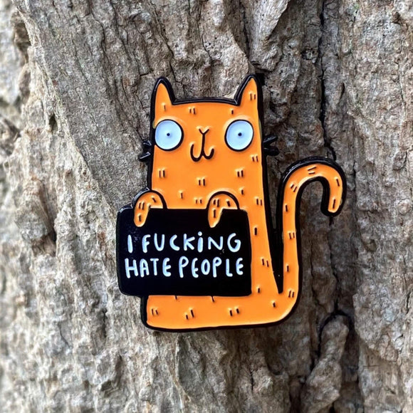 Close-up of the Katie Abey soft enamel pin depicting an orange cat holding a sign that says 'I Fucking Hate People'. The pin is positioned against the bark of a tree, highlighting its quirky design and contrast with the natural background.