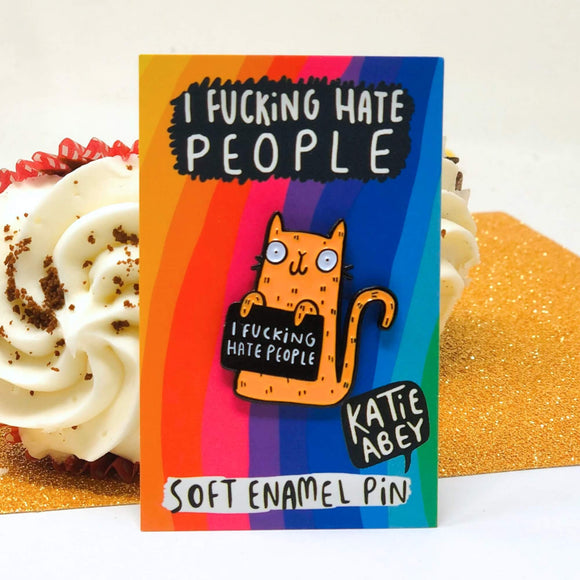 Katie Abey soft enamel pin featuring an orange cat holding a black sign that reads 'I Fucking Hate People'. The pin is displayed on a colourful backing card with a rainbow gradient background and the same phrase written at the top. The card also includes the artist's name, Katie Abey, in a speech bubble.